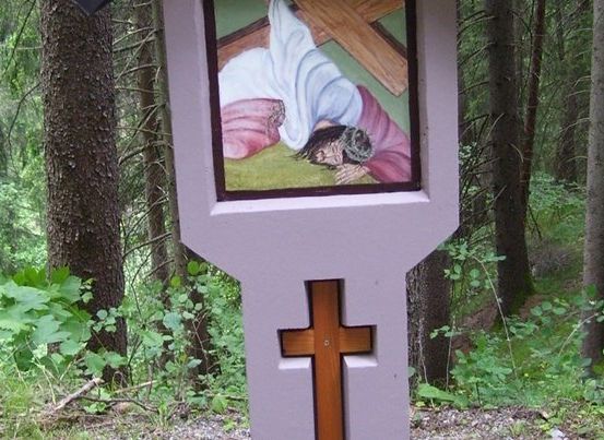 Ein hölzernes Kreuz mit einer gemalten Szene eines Mannes, der ein Kreuz auf dem Rücken trägt, steht in einem Wald.