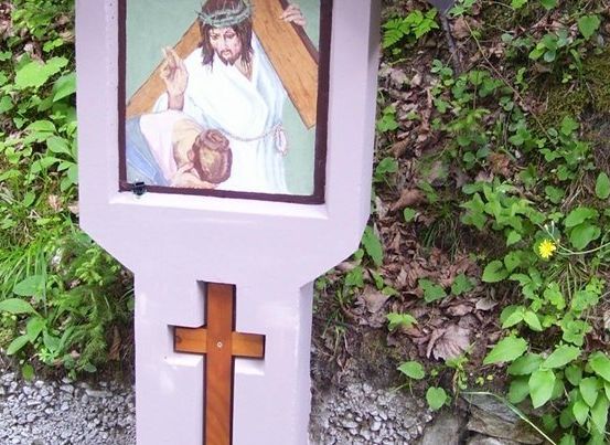 Ein hölzernes Kreuz auf einem lila Posten mit einem Gemälde von Jesus, der das Kreuz trägt, und einem Mann, der vor ihm kniet.