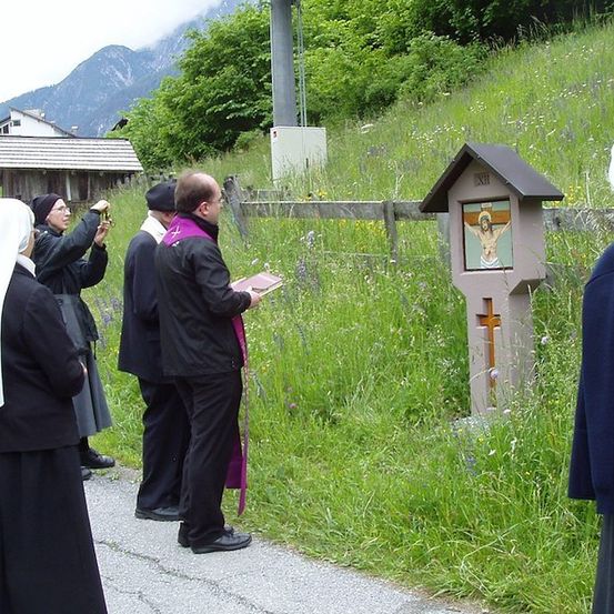 Eine Gruppe von Menschen, einschließlich eines Priesters, versammelt sich um ein kleines Heiligtum in einem Grasbereich. Eine Person macht ein Foto.