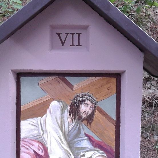 Eine kleine Kapelle mit einem Gemälde von Jesus, der das Kreuz trägt. Die Kapelle hat ein Dach und ist rosa gestrichen. Das Gemälde ist gerahmt und hat die Nummer VII darüber.