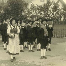 Ein altes Foto einer Marschkapelle in traditioneller Kleidung, die Instrumente hält und in einer Reihe geht.