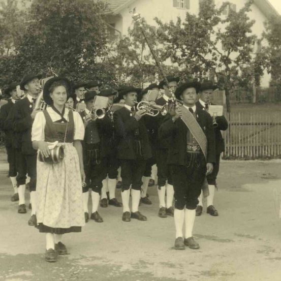 Ein altes Foto einer Marschkapelle in traditioneller Kleidung, die Instrumente hält und in einer Reihe geht.