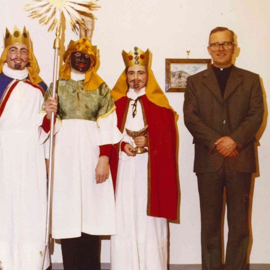 Vier Männer in traditionellen Kostümen, zwei mit Kronen, einer hält einen Stab und ein anderer einen Kelch, stehen vor einer weißen Wand mit einem gerahmten Bild.