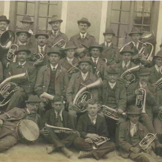 Ein altes Foto einer Blechbläserband, alle in Anzügen und Hüten gekleidet, halten verschiedene Blechblasinstrumente. Die Bandmitglieder sind in Reihen angeordnet, einige sitzen und andere stehen. Das Bild hat einen Sepiaton.
