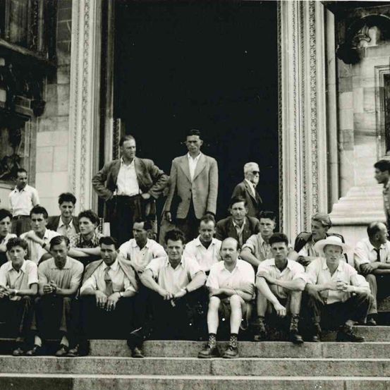 Eine Gruppe von Männern in formeller Kleidung, einige sitzend und andere stehend, posiert für ein Foto vor einem Gebäude.