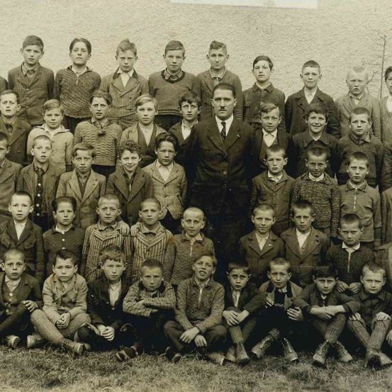Ein altes Schwarz-Weiß-Foto einer Gruppe junger Jungen und eines Mannes, möglicherweise ein Lehrer, vor einer Wand.