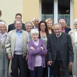 Eine Gruppe älterer Menschen posiert für ein Foto vor einem Gebäude. Sie alle lächeln und scheinen glücklich zu sein.