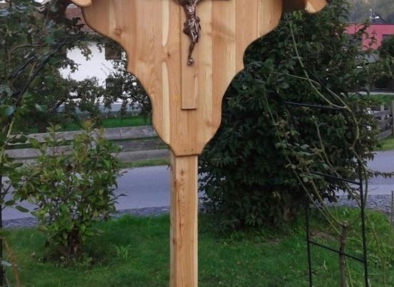 Ein hölzernes Kruzifix steht in einem Garten, mit einem Metallzaun und Grünpflanzen im Hintergrund.