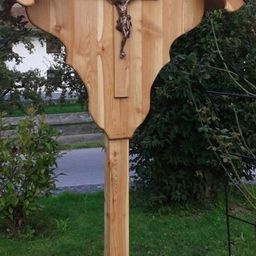 Ein hölzernes Kruzifix steht in einem Garten, mit einem Metallzaun und Grünpflanzen im Hintergrund.