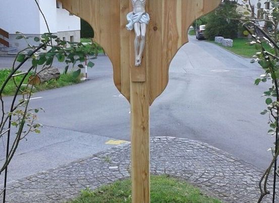 Ein hölzernes Kruzifix ist an einem hölzernen Pfosten am Straßenrand angebracht, umgeben von grünen Pflanzen.