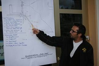 Ein Mann in einer schwarzen Jacke und mit Brille zeigt auf eine Tafel mit einem Diagramm und Text.