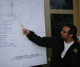 Ein Mann in einer schwarzen Jacke und mit Brille zeigt auf eine Tafel mit einem Diagramm und Text.