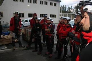 Eine Gruppe von Personen, die in rote und schwarze Sicherheitsausrüstung, Helme und Handschuhe gekleidet sind, bereitet sich möglicherweise auf eine Outdoor-Aktivität auf einem Parkplatz vor.