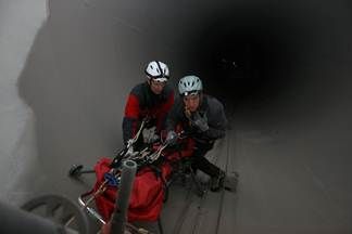 Zwei Personen mit Helmen und Schutzausrüstung arbeiten an einem rot-schwarzen Fahrzeug in einem Tunnel. Einer sitzt auf einem Wagen mit Rädern, während der andere in der Nähe steht.