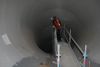 Ein Bauarbeiter mit Helm und Schutzausrüstung steht auf einer Gerüstkonstruktion in einem großen Tunnel. Die Tunnelwände sind unfertig, und ein kleines Stück weißes Papier ist an der Wand befestigt.