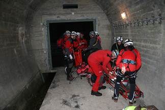 Eine Gruppe von Personen in roten Schutzanzügen und Helmen befindet sich in einem Tunnel und kümmert sich um jemanden auf einer Trage. Die Tunnelwände sind aus Beton und rechts befindet sich eine Lampe.