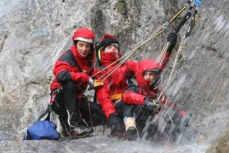 Drei Kletterer in roten Jacken und Handschuhen posieren für ein Foto beim Klettern an einem Wasserfall. Sie halten sich an Seilen und Gurten fest.