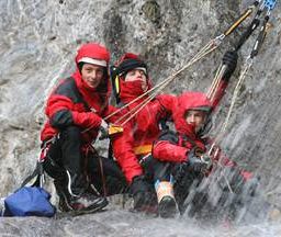 Drei Kletterer in roten Jacken und Handschuhen posieren für ein Foto beim Klettern an einem Wasserfall. Sie halten sich an Seilen und Gurten fest.