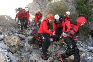 Eine Gruppe von Bergsteigern in roten Jacken und Helmen klettert einen felsigen Pfad hinauf, wobei eine Person von anderen unterstützt wird.
