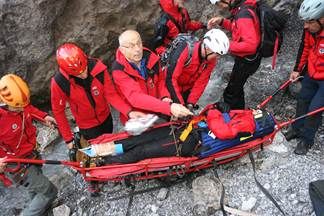 Eine Gruppe von Menschen in roten Jacken und Helmen trägt eine Trage mit einer Person darauf. Sie befinden sich auf einer felsigen Oberfläche, möglicherweise in einem Berggebiet.