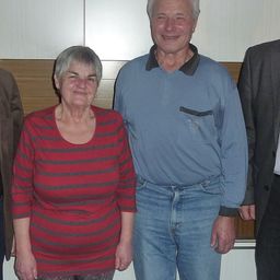 Vier Personen stehen zusammen und lächeln für ein Foto. Eine Frau in einem rot gestreiften Shirt steht zwischen zwei Männern. Der Mann links trägt einen braunen Mantel, während der Mann rechts einen Anzug und eine Krawatte trägt.
