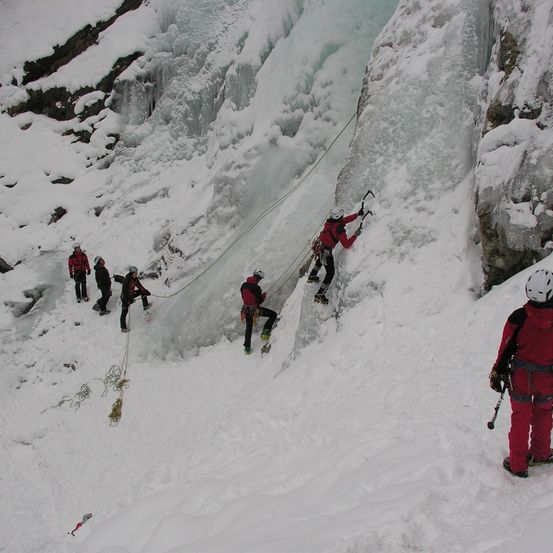 Eine Gruppe von Kletterern in roter Ausrüstung erklimmt einen verschneiten Berg und benutzt Seile und Eispickel.