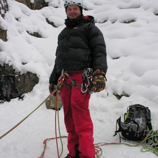 Ein Mann in einer schwarzen Jacke und roten Hosen steht auf schneebedecktem Gelände, hält Seile und Karabiner und hat zwei Rucksäcke im Hintergrund.