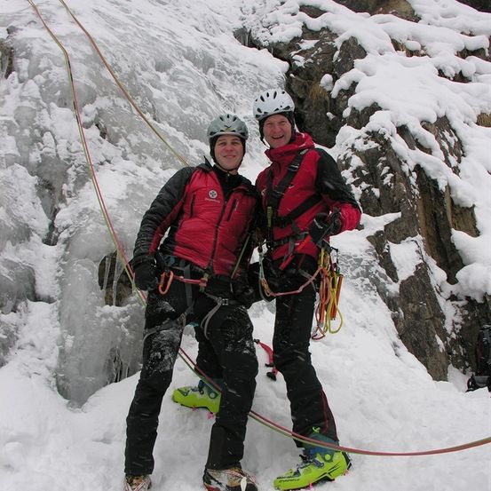 Zwei Kletterer in roten Jacken und Helmen stehen auf einem verschneiten Berg und lächeln für ein Foto. Sie sind mit Seilen und Kletterausrüstung ausgestattet.
