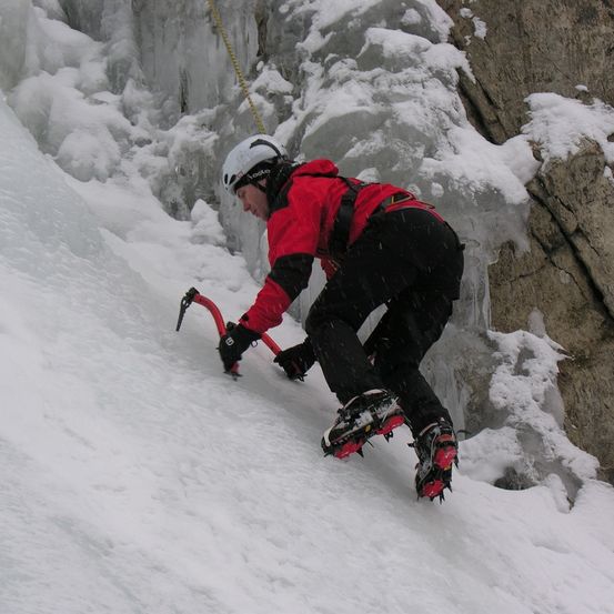 Ein Kletterer in einem roten Jacket und weißem Helm klettert an einer verschneiten Eiswand mit Steigeisen und Eispickeln hoch.