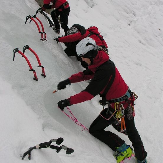 Eine Gruppe von Kletterern in roten Jacken und Helmen erklimmt einen vereisten Berg mit Eispickeln und Seilen.
