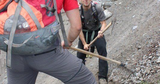 Zwei Männer arbeiten auf einem felsigen Pfad, einer hält eine Spitzhacke und der andere trägt einen Rucksack.