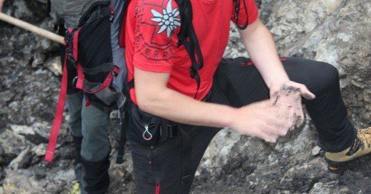 Eine Person in einem roten Shirt und schwarzen Hosen klettert einen felsigen Pfad hinauf und hält ein kleines Objekt in der Hand. Eine weitere Person in Grau steht hinter ihnen mit einem Rucksack.