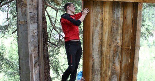 Ein Mann in einem roten Jackett klettert an einer Holzstruktur hoch, möglicherweise einem Baumhaus, während er sich an einer Holztür festhält.