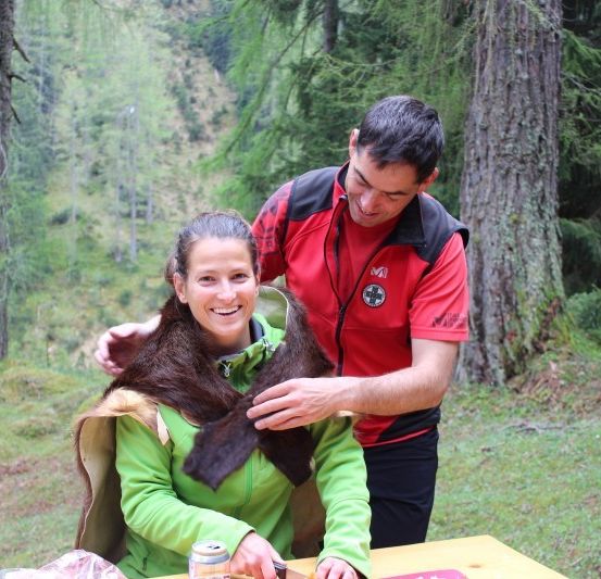 Ein Mann und eine Frau stehen in einem Wald, der Mann justiert einen Pelzschal um den Hals der Frau.