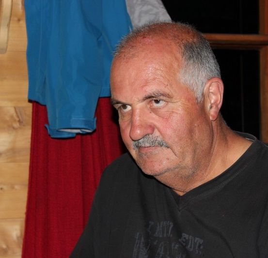 Ein älterer Mann mit einem Schnurrbart und grauen Haaren, der ein schwarzes T-Shirt trägt, sitzt in einem Raum mit Holzwänden und einem roten Tuch, das hinter ihm hängt.