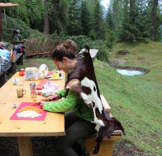 Eine Frau mit einem über die Schultern geworfenen Ziegenfell schneidet Essen auf einem Picknicktisch im Wald.