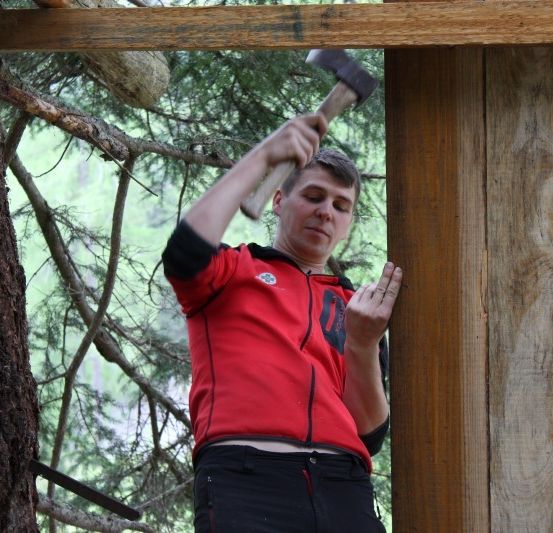 Ein Mann in einem roten Jackett benutzt einen Axthammer, um eine Holzstruktur in einem Wald zu schlagen.