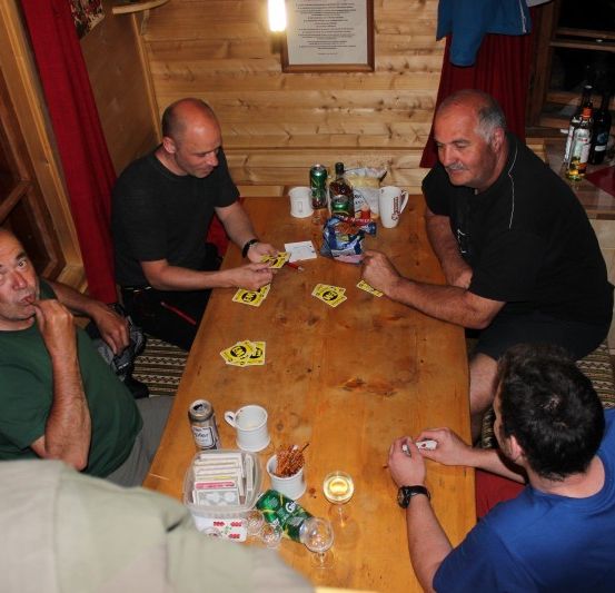 Vier Männer sitzen um einen Holztisch und spielen ein Kartenspiel. Ein Mann hält eine Zigarette und ein anderer eine Karte. Auf dem Tisch stehen Tassen, Flaschen und Snacks.
