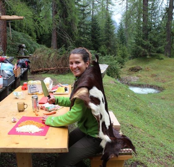 Eine Frau sitzt an einem Picknicktisch im Freien, trägt einen Pelzmantel und bereitet Essen zu.