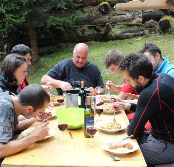 Eine Gruppe von Menschen sitzt um einen Tisch herum und isst und trinkt. Sie befinden sich in einer Außenumgebung mit Bäumen und einer Holzstruktur im Hintergrund.