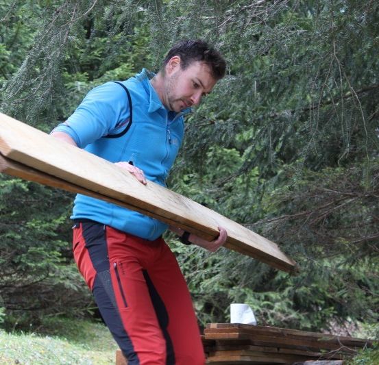 Ein Mann in einem blauen Jackett und roten Hosen trägt eine große Holzplanke im Freien. Er steht in einem Wald mit Bäumen im Hintergrund.
