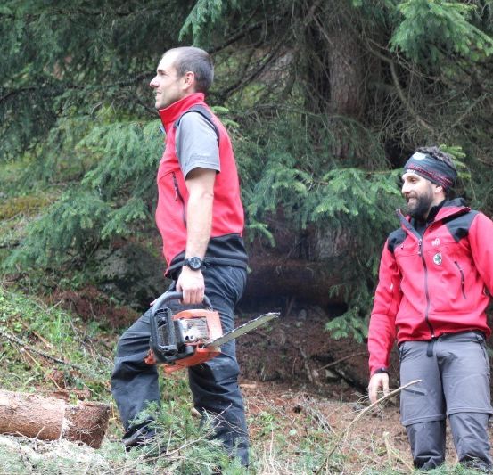 Zwei Männer in roten Jacken stehen in einem Wald, einer hält eine Kettensäge und der andere schaut ihn an.