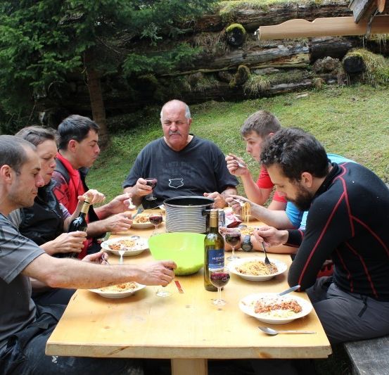 Eine Gruppe von Männern sitzt um einen Holztisch herum, isst Essen und trinkt Wein. Sie befinden sich in einem Grasbereich mit Bäumen und einer Holzstruktur im Hintergrund.