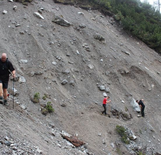 Eine Gruppe von Wanderern klettert einen felsigen Bergpfad hinauf. Ein Mann führt mit Trekkingstöcken, während andere hinter ihm herkommen.