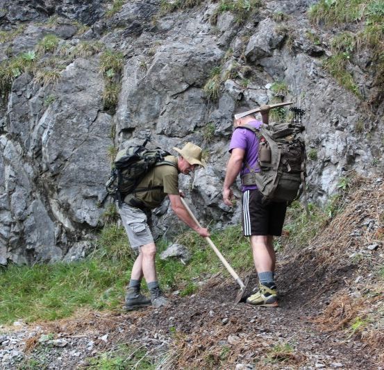Zwei Wanderer, einer mit einer Schaufel, räumen einen Pfad auf einer felsigen Bergseite.