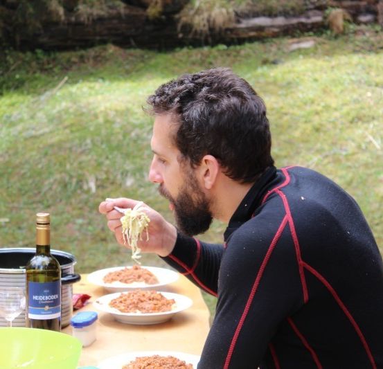 Ein Mann isst Spaghetti im Freien und sitzt an einem Tisch mit einer Flasche Wein und zwei Tellern Essen.