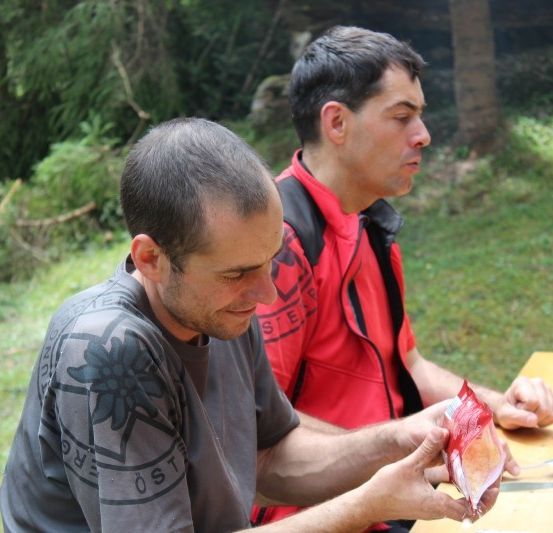 Zwei Männer sitzen an einem Picknicktisch in einer grasigen Gegend, einer öffnet eine rote Tüte, während der andere zusieht.