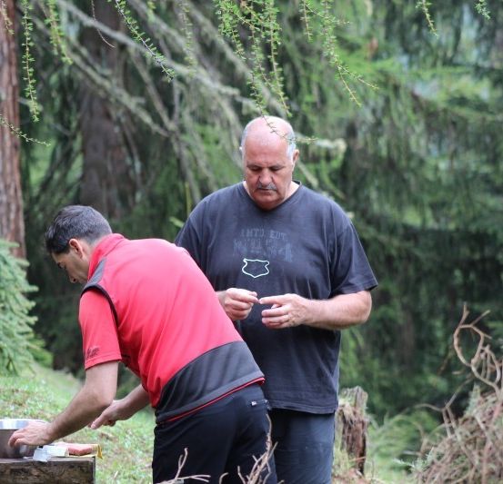 Zwei Männer sind im Freien in einem Wald. Einer gießt Wasser aus einem Metallbecher in eine Schüssel. Der andere Mann schaut etwas in seiner Hand an.