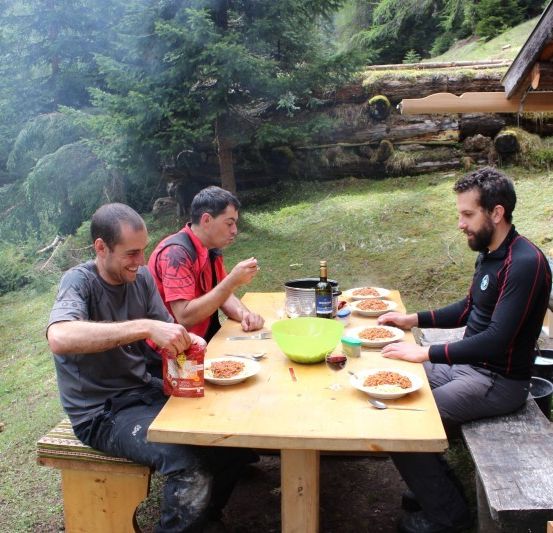 Drei Männer sitzen an einem Holztisch im Wald und genießen eine Mahlzeit. Ein Mann lächelt beim Essen, ein anderer hält eine Schüssel und der dritte schaut in die Kamera. Dahinter steht eine Holzbank und ein Tisch mit Flaschen und Essen.