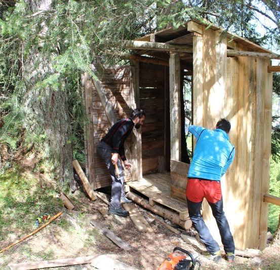 Zwei Männer arbeiten an einer Holzstruktur im Wald, einer hält einen Hammer und der andere steht neben einem Baum.